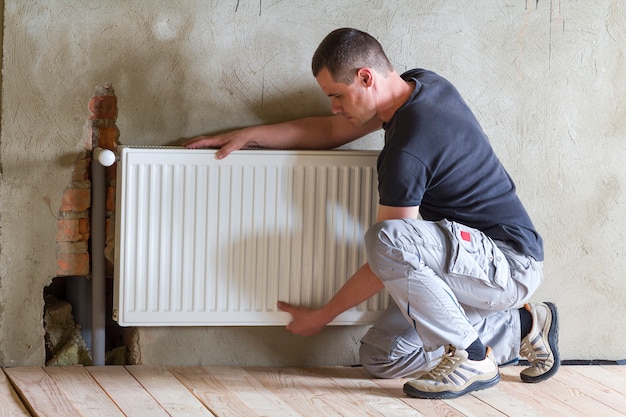 Jonge knappe professionele loodgieter werknemer verwarming radiator installeren in een lege ruimte van een nieuw gebouwde appartement of huis. Concept van constructie, onderhoud en reparatie.