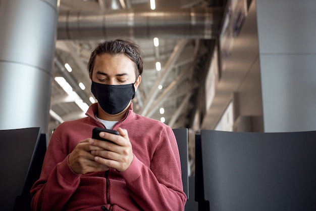 Jonge knappe reiziger in een masker in de lounge van de luchthaventerminal met smartphone-app in openbare wifi-ruimte, messaging