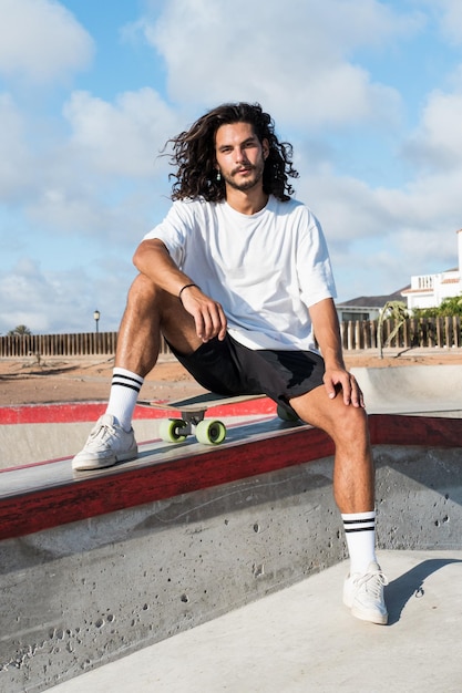 Foto jonge knappe skater zittend op zijn skateboard in het skatepark