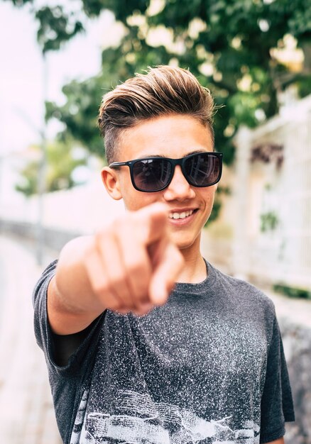 Jonge knappe tiener portret wijzende vinger naar jou en glimlach - blonde blanke jongen outdoor park vrijetijdsbesteding met zonnebril en gelukkige uitdrukking