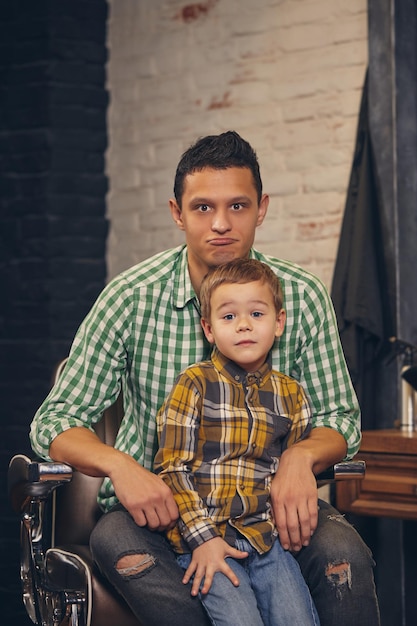 Jonge knappe vader en zijn stijlvolle zoontje bij kapperszaak wachten op kapper. De vader zit in de stoel en de zoon staat vlakbij