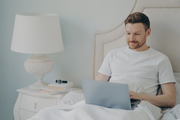 Jonge knappe vrolijke glimlachende man freelancer in casual pyjama-outfit die 's ochtends in bed zit en thuis op afstand op laptop werkt terwijl hij in een gezellig bed rust. Freelance concept