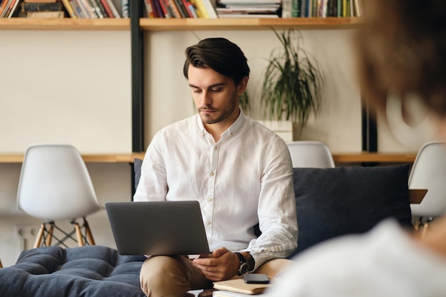Jonge knappe zakenman zittend op de bank bedachtzaam bezig met laptop in moderne coworking space