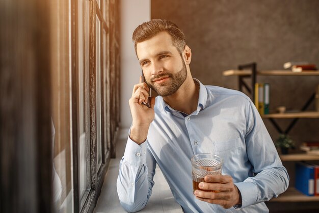 Jonge knappe zakenmantribune bij venster en bespreking op telefoon in zijn eigen bureau. Hij houdt een glas whisky in de hand en kijkt naar buiten. Zelfverzekerde aardige jongeman.