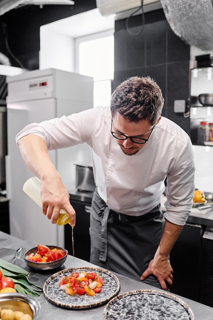 Jonge kok in schort die olijfolie in salade op bord toevoegt voor het serveren terwijl hij in de keuken staat