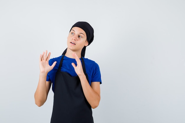 Jonge kok man met handpalmen in overgave gebaar in t-shirt, schort en bang kijken. vooraanzicht.