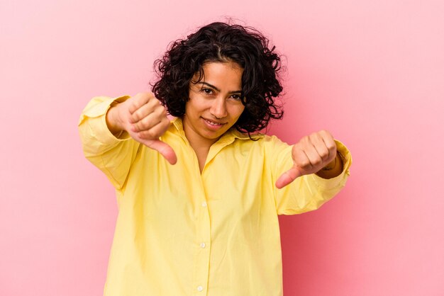 Jonge krullende Latijnse vrouw die op roze achtergrond wordt geïsoleerd die duim neer toont en afkeer uitdrukt.