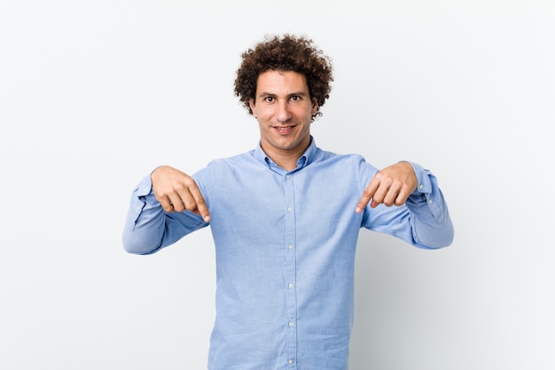 Jonge krullende volwassen man met een elegant shirt wijst naar beneden met vingers, positief gevoel.