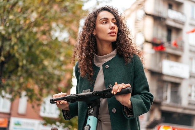 Jonge krullende vrouw met elektrische scooter in de stad