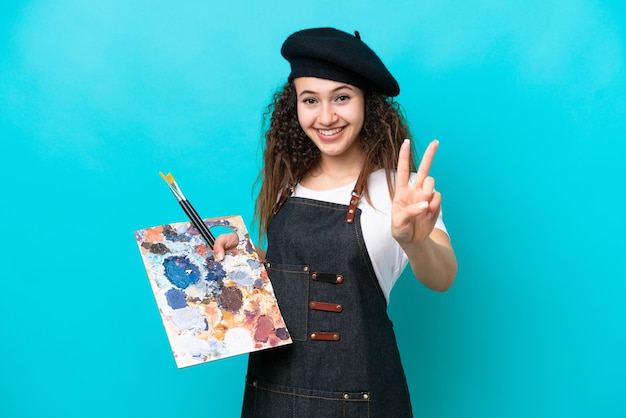 Jonge kunstenaar Arabische vrouw met een palet geïsoleerd op blauwe achtergrond glimlachend en overwinningsteken tonen