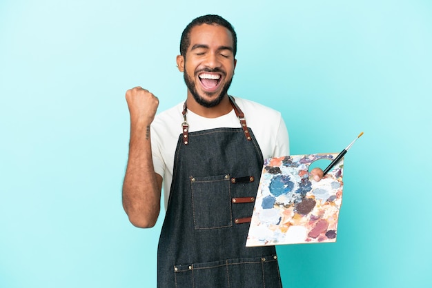 Jonge kunstenaar Latijns-man met een palet geïsoleerd op blauwe achtergrond vieren een overwinning