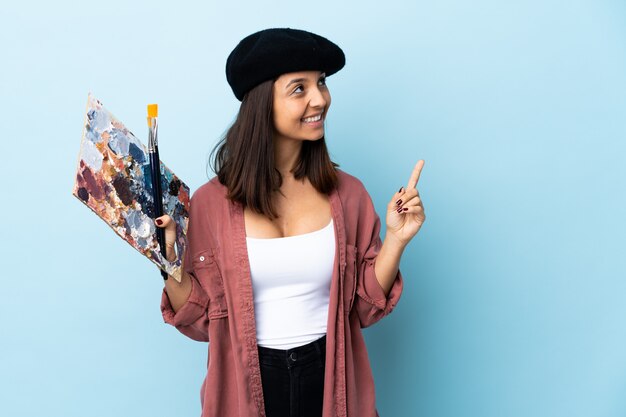 Foto jonge kunstenaarsvrouw die een palet over geïsoleerde blauwe achtergrond houdt die een groot idee benadrukt