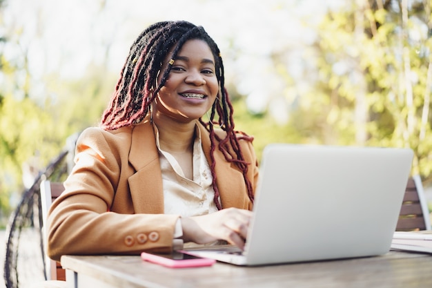 Jonge lachende Afro-Amerikaanse vrouw die aan de tafel zit in een straatcafé en een laptop gebruikt?