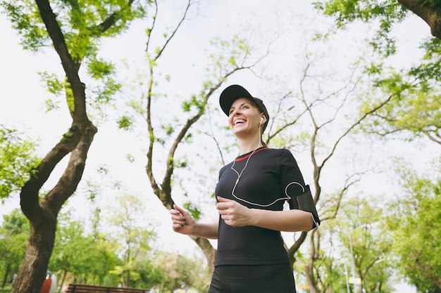 Jonge lachende atletische brunette vrouw in zwart uniform en pet met koptelefoon training sport doen, hardlopen en luisteren naar muziek op pad in stadspark buitenshuis