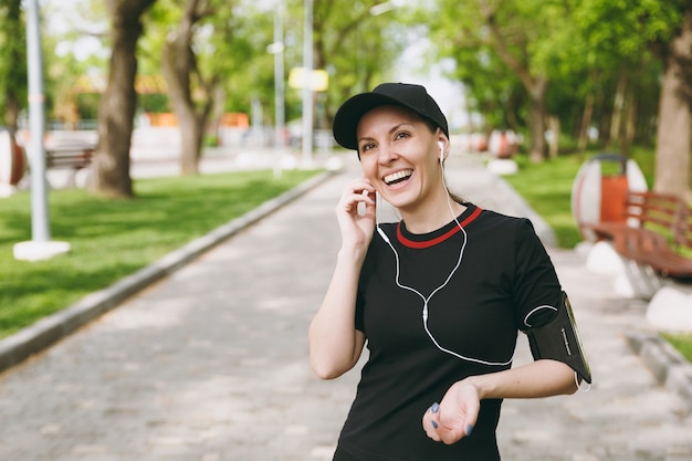 Jonge lachende atletische mooie brunette vrouw in zwart uniform en pet met koptelefoon luisteren naar muziek, hand in de buurt van oor houden op training in stadspark buitenshuis