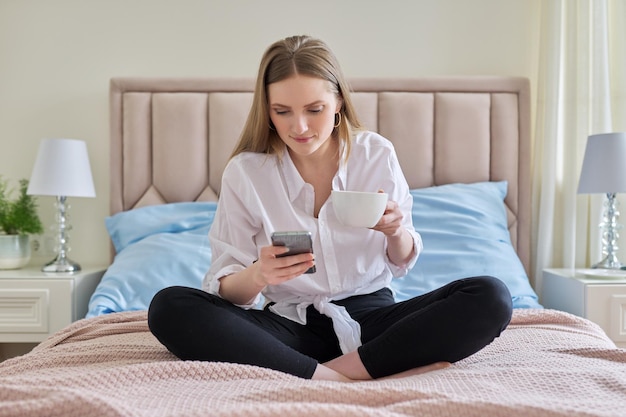 Jonge lachende blonde met kopje koffie en smartphone zittend in bed