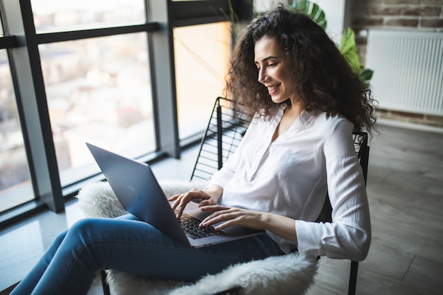 Jonge lachende brunette meisje zit op moderne stoel bij het raam in lichte, gezellige kamer thuis bezig met laptop in een ontspannen sfeer