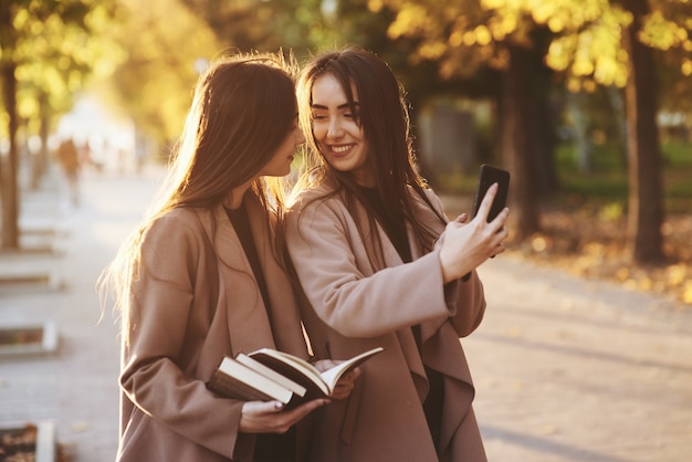 Jonge lachende brunette tweeling meisjes kijken elkaar en nemen selfie met zwarte telefoon, terwijl een van hen boeken vasthoudt, jas draagt, staande op herfst zonnige park steegje op onscherpe achtergrond.