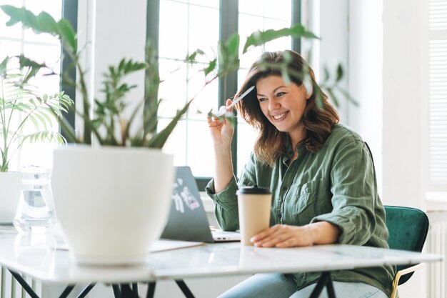 Jonge lachende brunette vrouw plus size werken op laptop op tafel met kamerplant in het lichte moderne kantoor