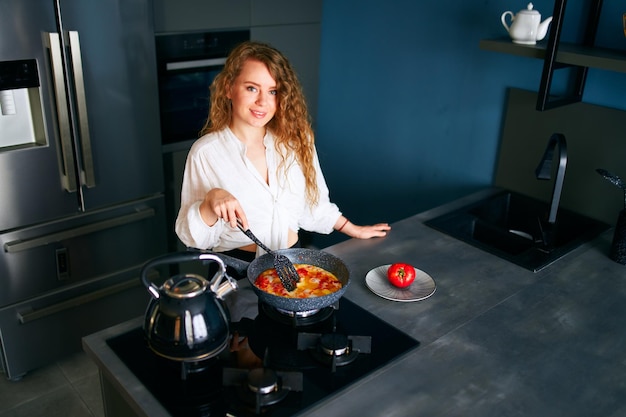 Jonge lachende gekrulde blanke vrouw kookt ontbijt op een koekenpan jonge volwassen vrouw making
