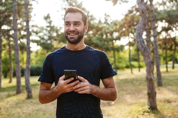 jonge lachende man in sportkleding met behulp van mobiele telefoon tijdens het sporten in zonnig groen park