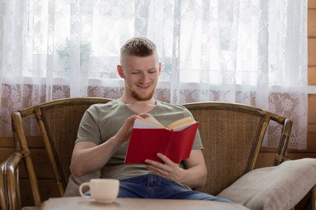 Jonge lachende man leesboek met rode kaft op rieten bank in houten huis van het platteland
