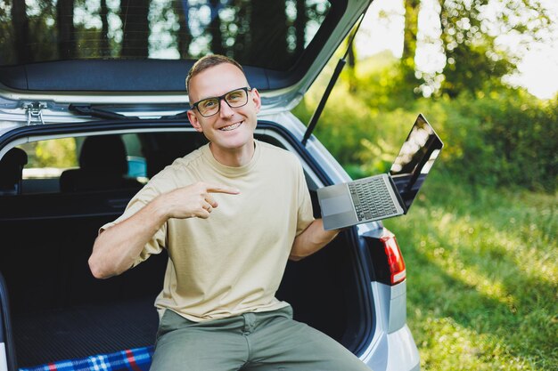 Jonge lachende man met bril freelancer werkt op een laptop terwijl hij in de kofferbak van een auto zit Werk op afstand in de natuur