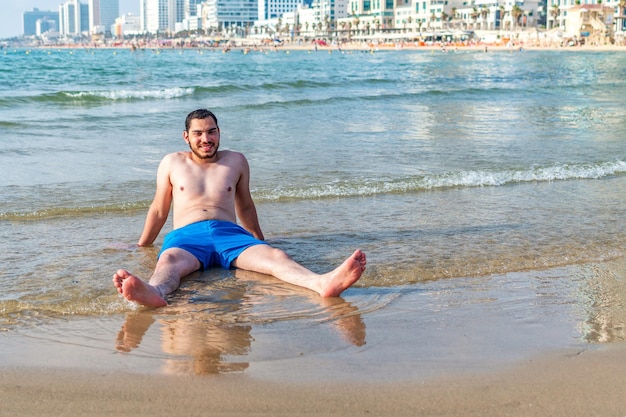 Jonge lachende man zittend op een strand camera kijken