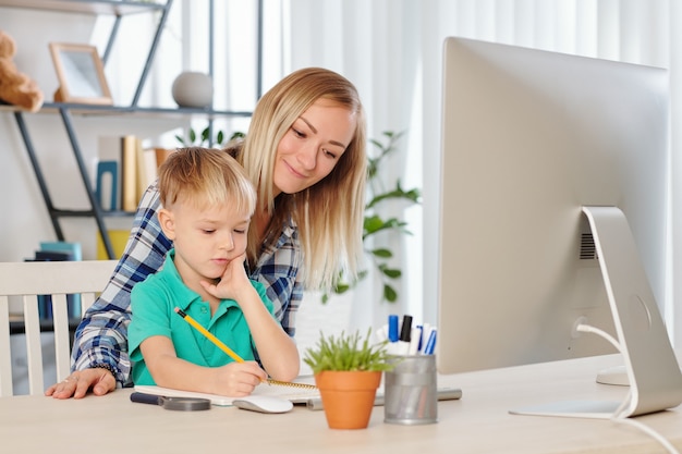 Jonge lachende moeder die haar zoontje controleert en huiswerk doet aan zijn bureau