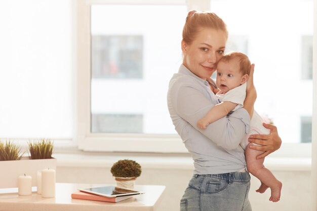 Jonge lachende moeder in blauw shirt en spijkerbroek en haar baby die thuis voor het raam staat