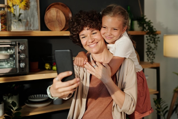 Jonge lachende moeder met smartphone die selfie neemt met gelukkige dochter