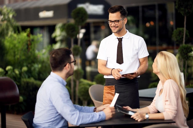 Jonge lachende ober die communiceert met gasten terwijl hij ze serveert in een restaurant