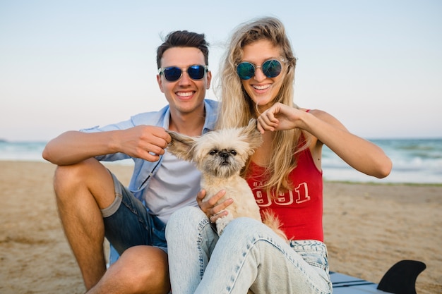 Jonge lachende paar plezier op het strand zittend op zand met surfplanken spelen met hond