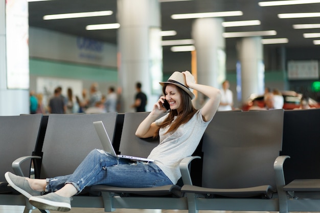 Jonge lachende reiziger toeristische vrouw die op laptop werkt, praat op mobiele telefoon, belt vriend op luchthaven