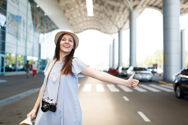 Jonge lachende reiziger toeristische vrouw in hoed met retro vintage fotocamera met papieren kaart, vangt taxi op internationale luchthaven