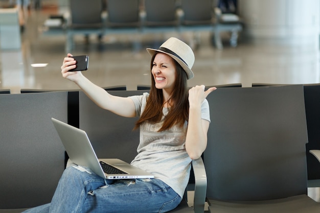 Jonge lachende reizigerstoeristenvrouw die aan laptop werkt die selfie op mobiele telefoon doet die in ontvangsthal bij internationale luchthaven wacht