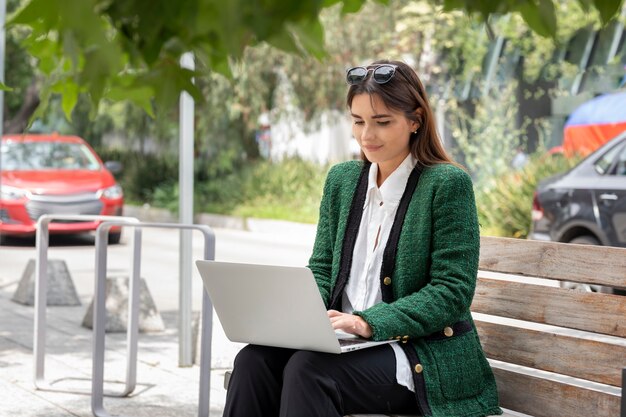 Jonge lachende vrouw aan het werk met haar laptop vanaf de straat zittend op een houten bankje