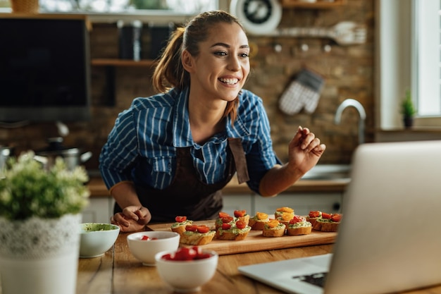 Jonge lachende vrouw die eten klaarmaakt in de keuken terwijl ze live streamt via laptop in de keuken