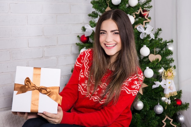 Jonge lachende vrouw die geschenkdoos opent in de buurt van versierde kerstboom