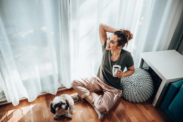 Jonge lachende vrouw genieten van koffie met haar Shih Tzu-hond in de gezellige ochtend thuis.