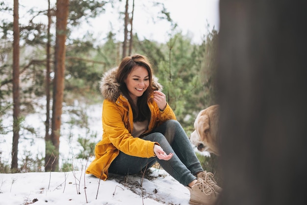 Jonge lachende vrouw in gele jas met grote vriendelijke witte hond Labrador wandelen in het winterbos