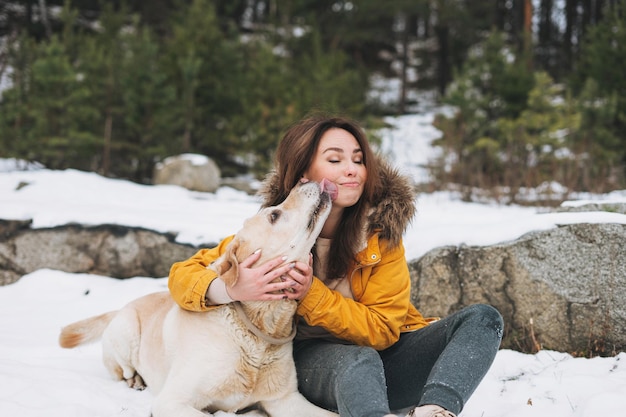 Jonge lachende vrouw in gele jas met grote vriendelijke witte hond Labrador wandelen in het winterbos