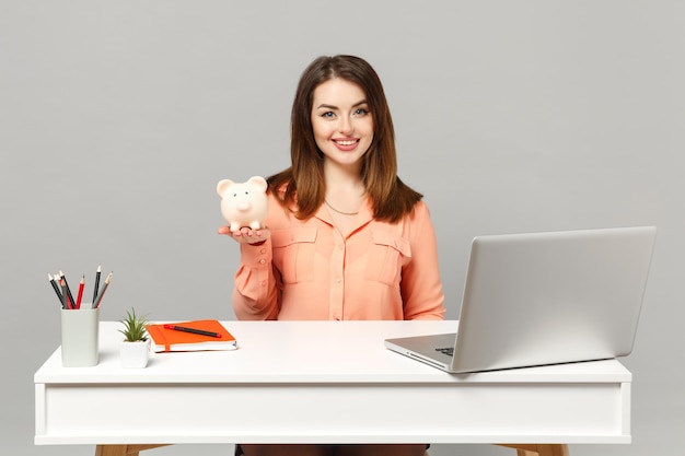 Jonge lachende vrouw in pastel casual kleding houdt spaarvarken zit werk aan witte bureau met pc-laptop geïsoleerd op een grijze achtergrond. Prestatie zakelijke carrière levensstijl concept. Bespotten kopie ruimte.