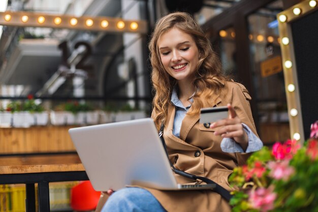 Jonge lachende vrouw in trenchcoat creditcard in de hand houden gelukkig bezig met laptop terwijl tijd buiten doorbrengen op gezellige café-terras