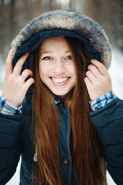 Jonge lachende vrouw in winterkleren met kap staande in winter woud, lachen, camera kijken. Winterlandschap op de achtergrond.