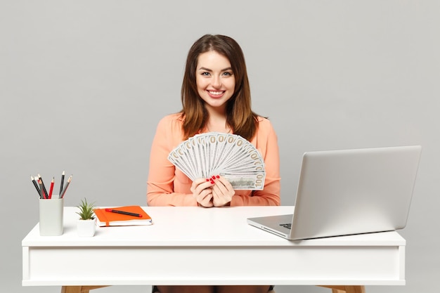 Jonge lachende vrouw met fan van contant geld in dollarbankbiljetten, zitten en werken aan bureau met pc-laptop geïsoleerd op een grijze achtergrond. Prestatie zakelijke carrière levensstijl concept. Bespotten kopie ruimte.