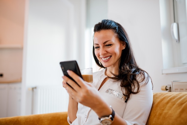 Jonge lachende vrouw ontspannen op de sofa met smartphone en warme drank.