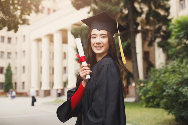 Jonge lachende vrouw op haar afstudeerdag in universiteit met diploma. onderwijs, kwalificatie en toga concept.