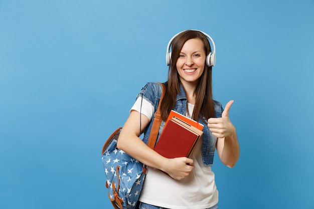 Jonge lachende vrouw student in denim kleding met rugzak, koptelefoon luisteren muziek, schoolboeken met duim opdagen geïsoleerd op blauwe achtergrond. Onderwijs in middelbare school hogeschool.