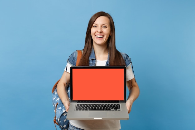 Jonge lachende vrouw student in denim kleding met rugzak met laptop pc-computer met leeg zwart leeg scherm geïsoleerd op blauwe achtergrond. Onderwijs op de universiteit. Kopieer ruimte voor advertentie.
